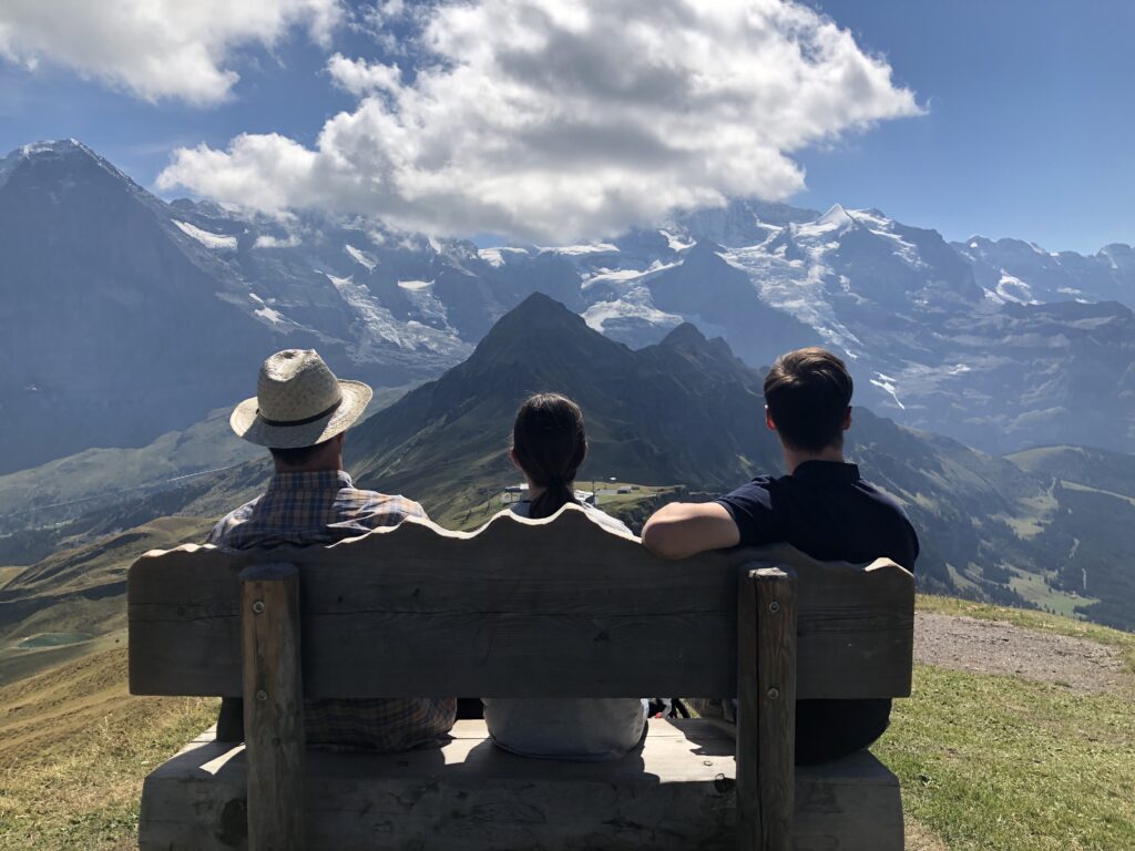 Vater sitzt mit seinen beiden Kindern auf einer Bank in den Schweizer Bergen und genießt die wunderschöne Aussicht