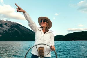 Frau im mittleren Alter steht am Ruder eines Bootes. Sie steuert über den See und sieht sehr glücklich aus. Dank Health Coaching füh lt sie sich gesund und attraktiv