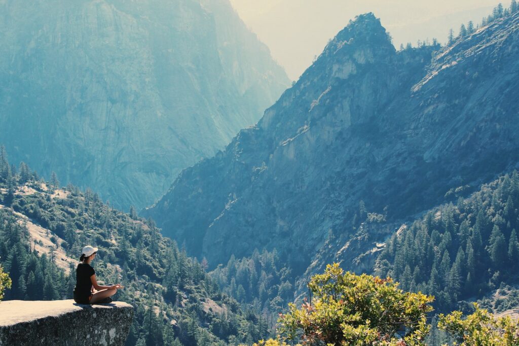 Junge Frau in den Bergen. Sie meditiert und ist sehr entspannt. Die blaue Farbe der Berge wirkt so beruhigend wie ein blaues Rainfarnöl