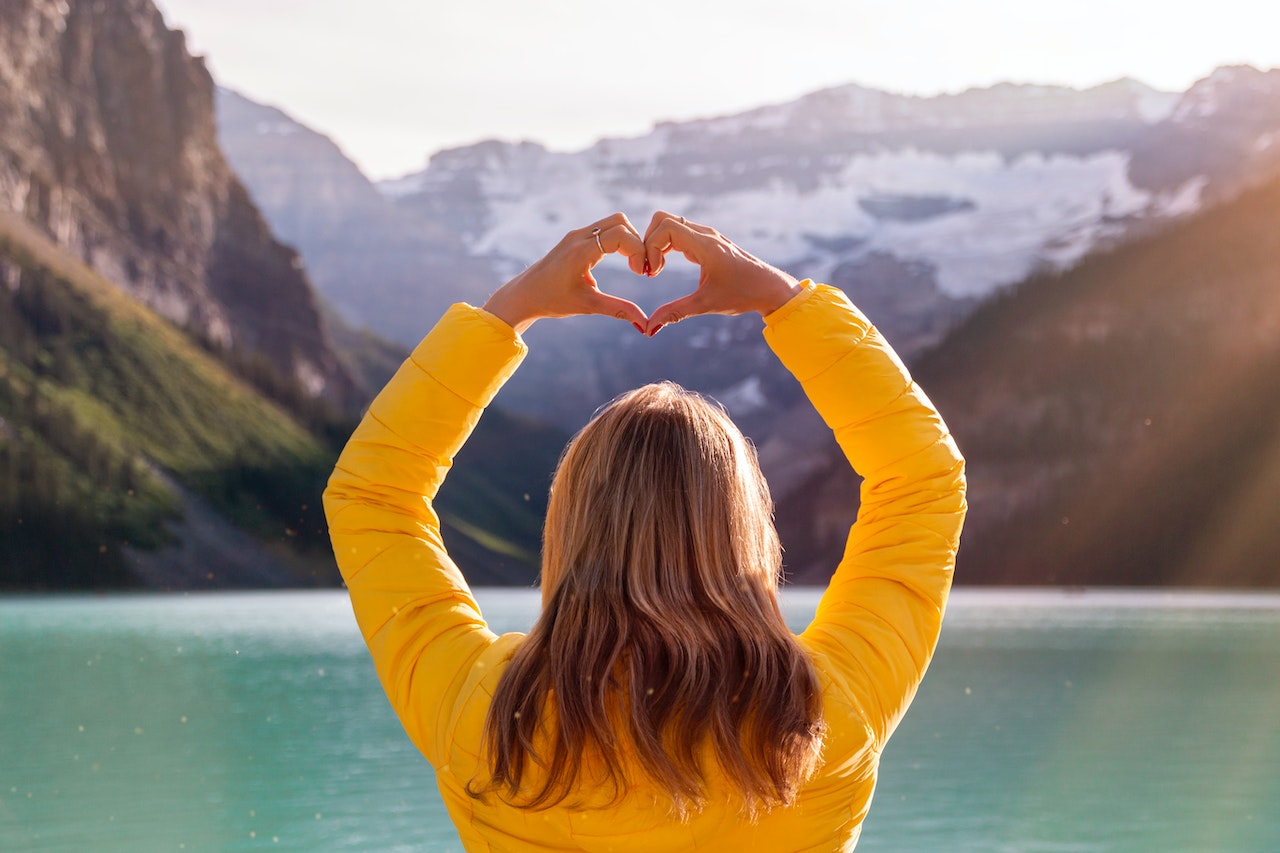 Frau über 40 mit gelber Jacke steht vor einem Bergsee und hebt die Arme. Sie formt ihre Händen zu einem Herz. Ihre Haltung zeigt, dass sie eine selbstbewusste, starke und gesunde Frau ist
