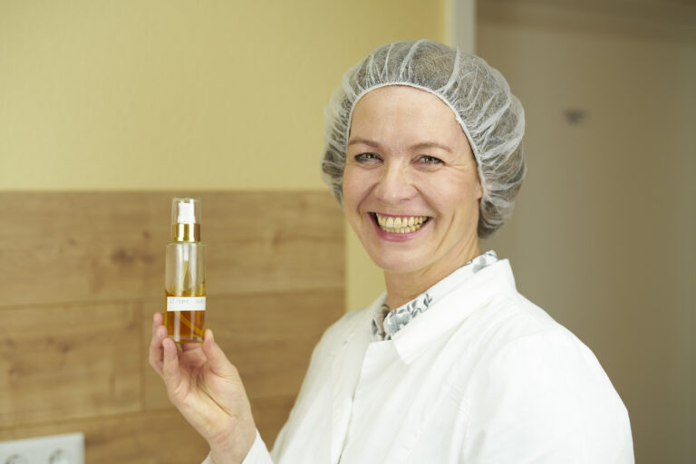 Naturkosmetik Entwicklerin Birgit Henttzler-Liepe in ihrem Labor. Sie trägt ihren Laborkittel und Haube.In ihrer hand hält sie eines der geprüften Gesichtsseren. Ihrer Erfahrung nach ist nicht einfach und es ein langer Weg, Naturkosmetik selber zu machen